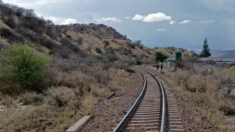 A single railway line running between overgrown heaps of historic mine waste, with a water tower and buildings ahead.